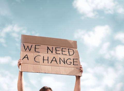 A person holds up a sign made out of cardboard which reads: "WE NEED A CHANGE."