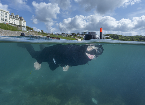 A man wearing a wetsuit is visible snorkelling underwater, with buildings visible on the horizon behind him