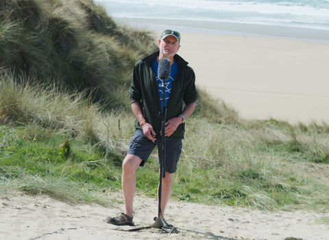 A white male stands in the marraum grass at Penhale Dunes nature reserve, in front of a microphone