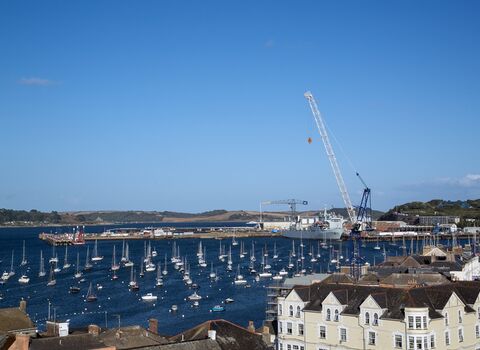 View across Falmouth Harbour showing numerous moored boats, a dockside crane, and waterside buildings under a clear blue sky.